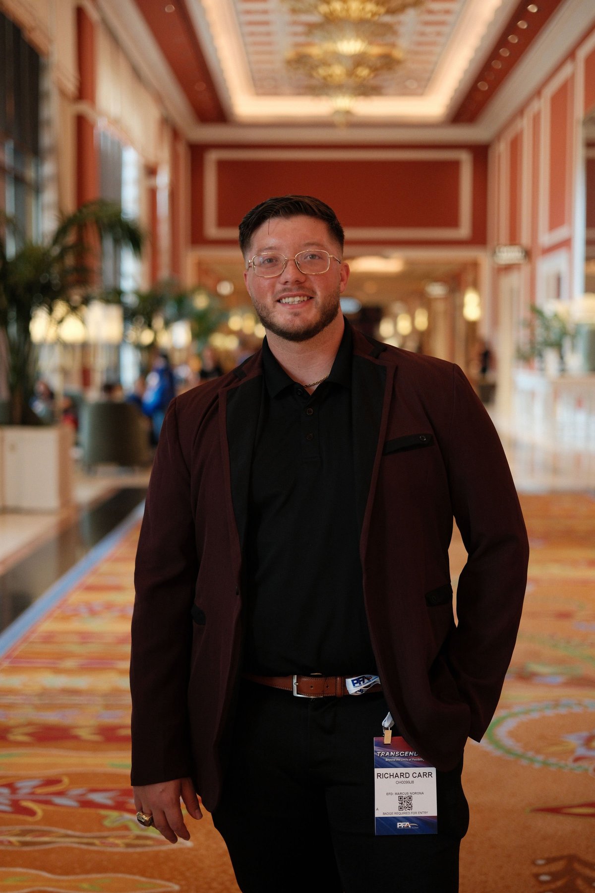 Ricky Randolph Carr standing in an elegant corridor, wearing a maroon blazer with a badge