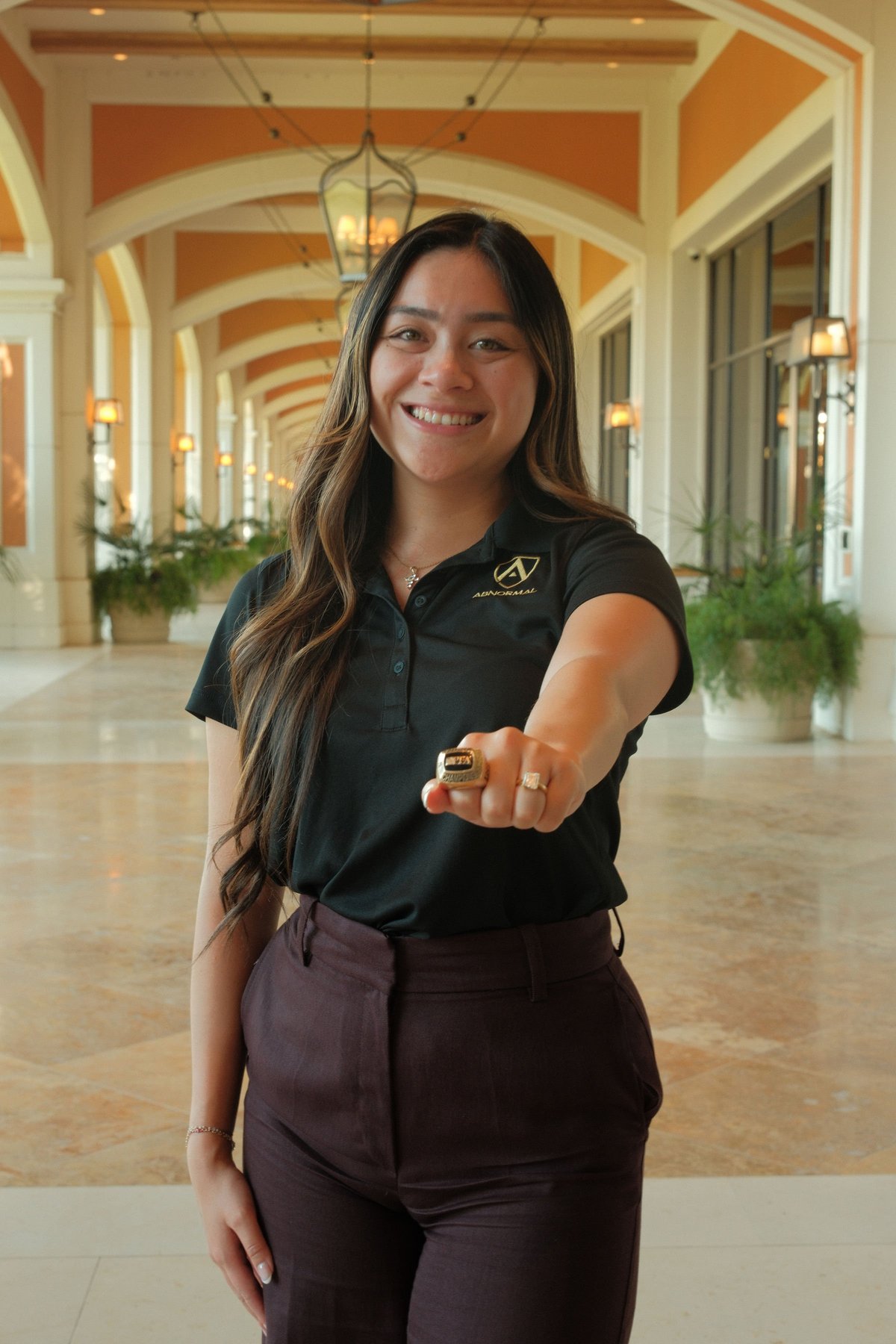 Caitlin Roque smiling in a black polo shirt, pointing toward the camera in an elegant hallway framed by orange and cream arches